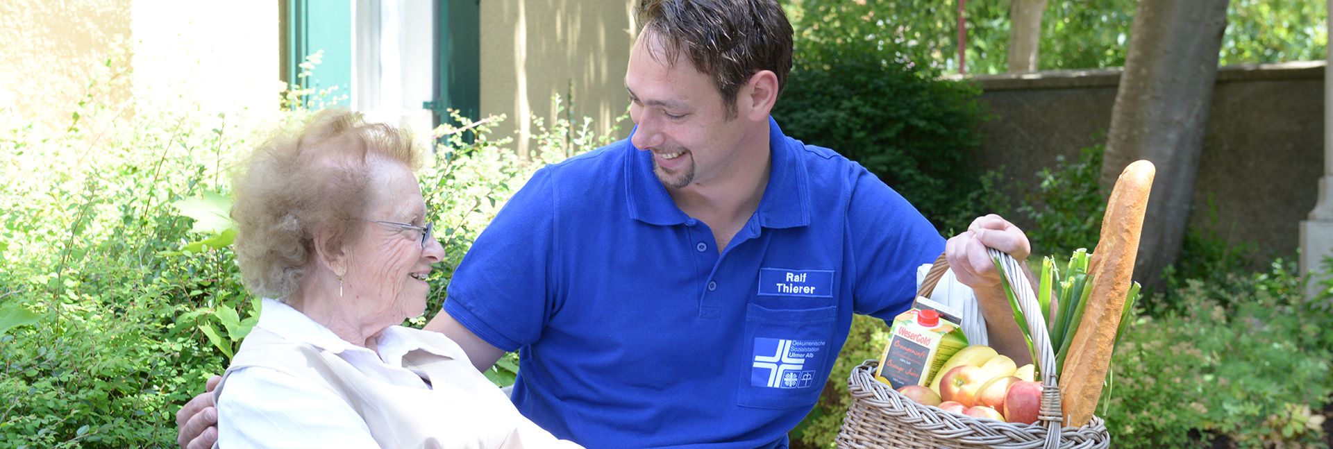 Mitarbeiter im blauen Polo übergibt einer Seniorin Einkaufskorb mit Obst