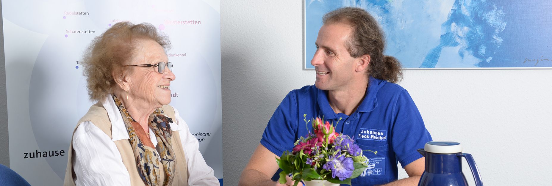 Ältere Frau im Gespräch mit lächelnder Pflegekraft am Tisch